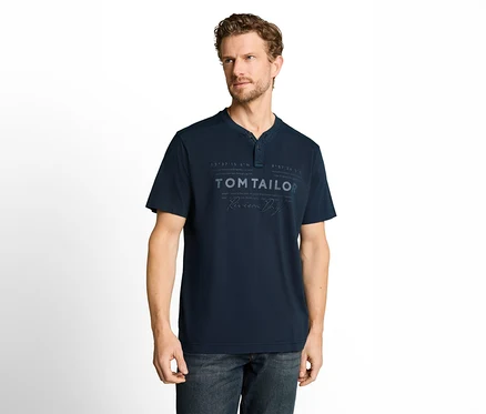 Göndör barna hajú, szakállas férfi sötétkék, rövid ujjú Henley nyakú pólót visel "TOM TAILOR" felirattal és sötét farmernadrágot. Fehér háttér előtt áll, jobbra néz.