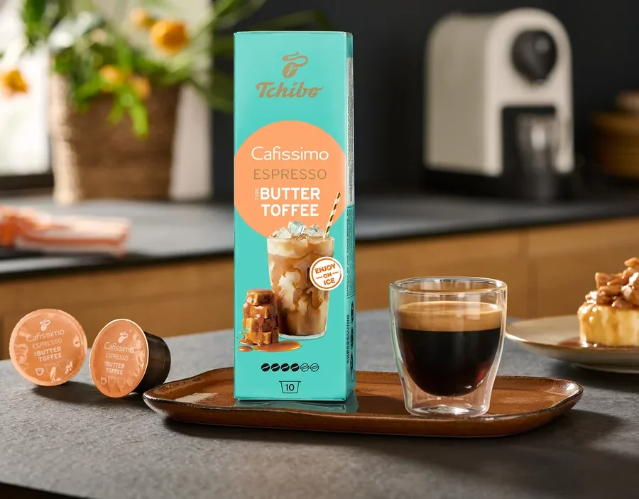 Egy csomag Cafissimo Flavoured Espresso – Buttertoffee és egy Cafissimo pure+, white kávéfőző áll az asztalon egy csésze kávé mellett.