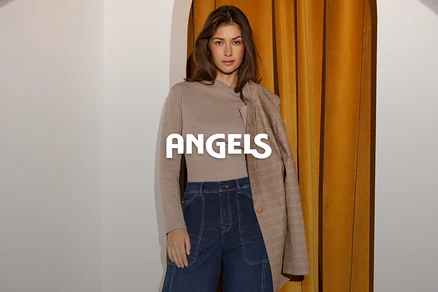 Egy nő bézs hosszú ujjú felsőben és sötétkék farmerben, barna kockás blézerrel a vállán, fehér fal és arany függöny előtt áll. Az ANGELS szó látható a képen.