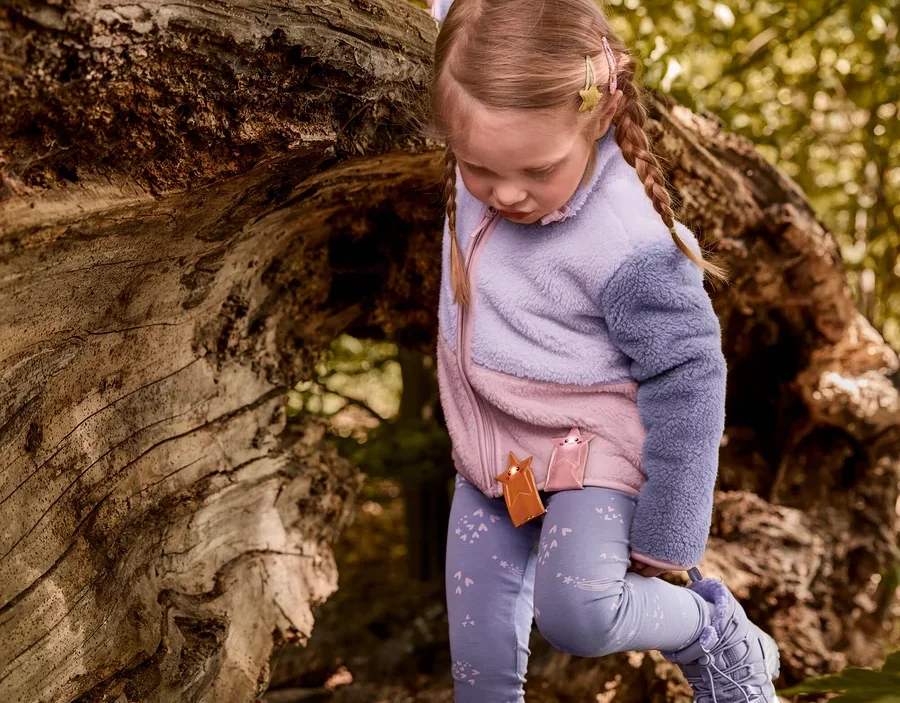Egy kislány lila-rózsaszín polár dzsekiben és lila leggingsben mászik egy kidőlt fatörzsön. Lila gyerek termobakancsot visel.