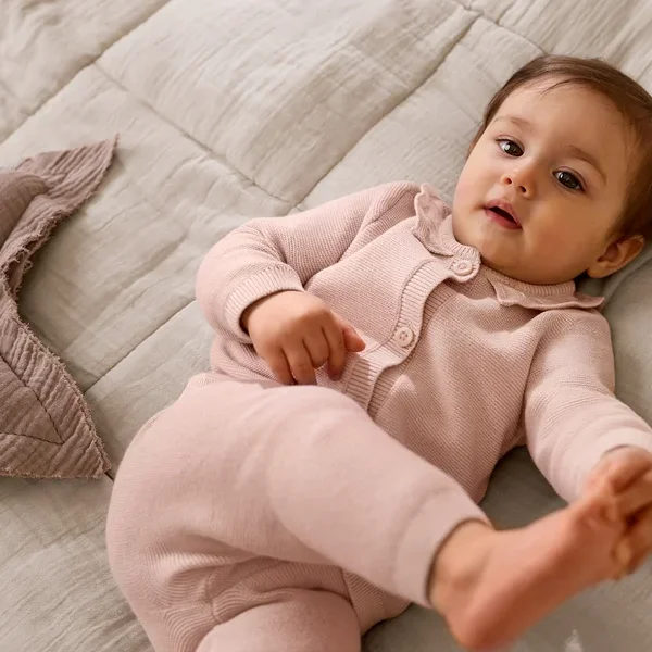 Egy rózsaszín kötött kardigánban és hozzá illő nadrágban lévő baba fekszik egy világosszürke, texturált takarón, felfelé nézve. A baba mellett egy rózsaszín csillag alakú párna van.