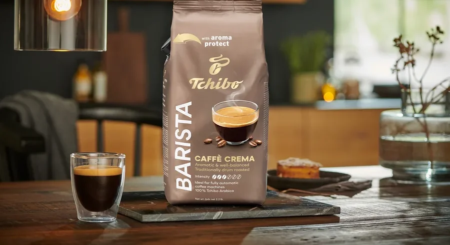Egy csomag BARISTA Caffè Crema - 1x1 kg szemes kávé egy asztalon áll egy pohár kávé mellett.