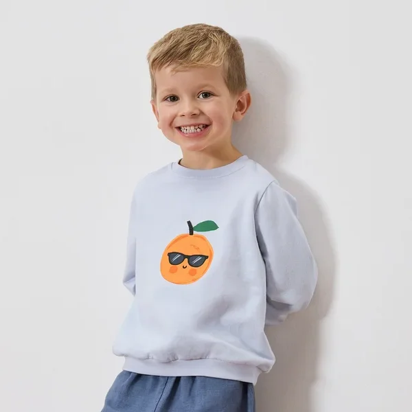 Lächelnder Junge trägt hellblaues Sweatshirt mit einer Orange mit Sonnenbrille darauf.