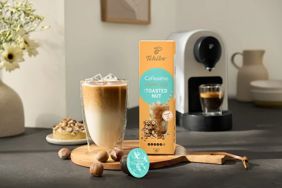Egy pohár jeges kávé áll a Cafissimo Flavoured Espresso – Toasted Nut csomag és egy Cafissimo pure+, white kávéfőző mellett.