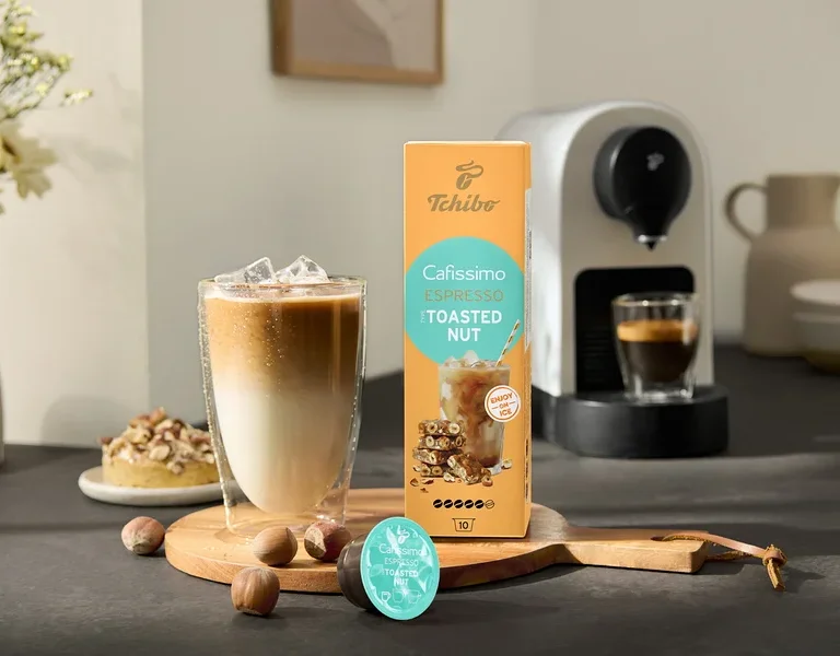 Egy pohár jeges kávé áll a Cafissimo Flavoured Espresso – Toasted Nut csomag és egy Cafissimo pure+, white kávéfőző mellett.