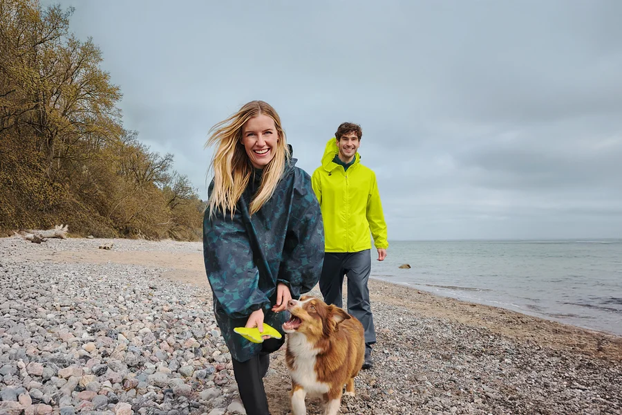 Eine lächelnde Frau in einem dunklen Regenponcho hält einen gelben Frisbee, während sie mit einem braun-weißen Hund an einem Kiesstrand spaziert. Ein lächelnder Mann in einer gelben Regenjacke geht hinter ihnen, mit Meer und bewölktem Himmel im Hintergrund.