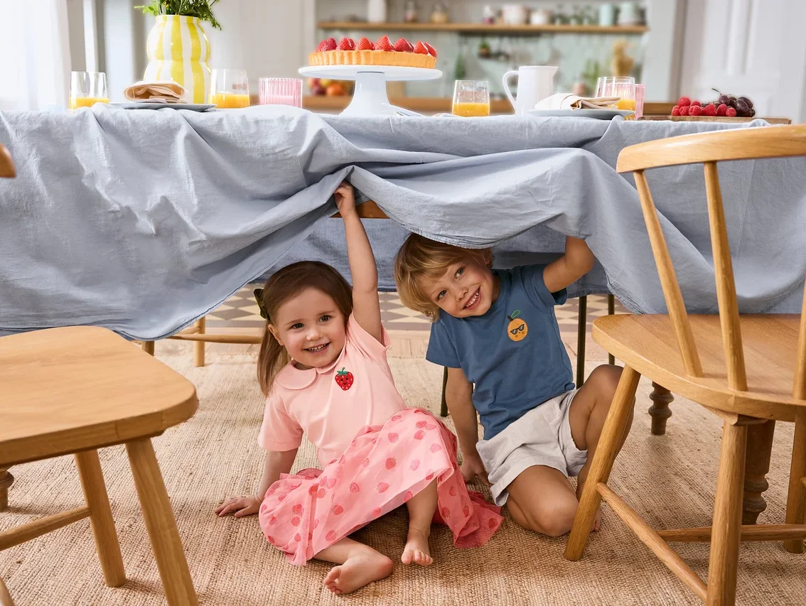 Két gyerek ül egy világoskék terítővel letakart asztal alatt. A lány Kinder-Tüllrock szoknyát és 2 Kinder-T-Shirts pólót visel. A fiú Kinder-Shorts rövidnadrágot visel.