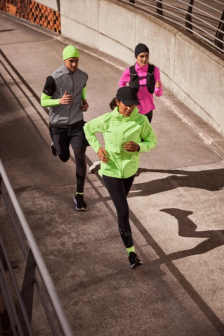 Három futó, két nő és egy férfi, fut egy betonúton. Az elöl lévő nő fekete sportsapkát, neonlime futódzsekit, fekete »ActivePower« sportleggingset és neon futózoknit visel. A középső férfi neonlime termosapkát, fényvisszaverő futómellényt fekete férfi sportfelső és futó karmelegítő felett, fekete férfi sport rövidnadrágot termo sportnadrág felett és neon futózoknit visel. A hátul lévő nő fekete termosapkát, rózsaszín női funkcionális termofelsőt és futómellényt visel.