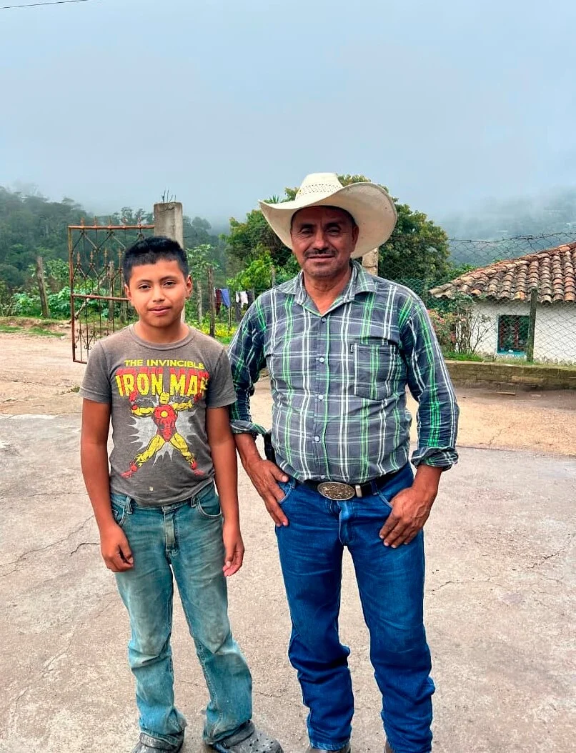 Egy felnőtt férfi cowboykalapban és egy fiú Iron Man pólóban állnak egymás mellett a szabadban. A férfi kockás inget és farmert visel, míg a fiú világoskék farmert. Burkolt felületen állnak, háttérben vidéki tájjal és egy házzal.