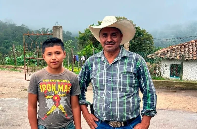 Egy felnőtt férfi cowboykalapban és egy fiú Iron Man pólóban állnak egymás mellett a szabadban. A férfi kockás inget és farmert visel, míg a fiú világoskék farmert. Burkolt felületen állnak, háttérben vidéki tájjal és egy házzal.