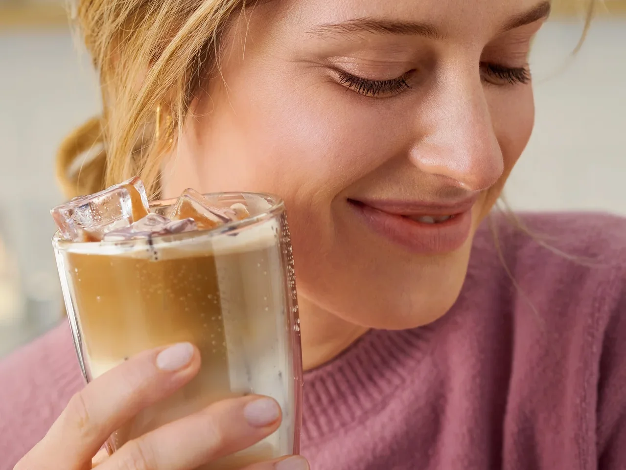 Nő tart egy poharat egy italral, ami jégkockákat tartalmaz. A háttérben egy fehér Cafissimo pure+ látható.
