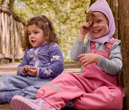 Két gyerek ül egy fa ösvényen. Az egyik gyerek lila gyerek termobakancsot és lila unikornisos felsőt visel. A másik gyerek rózsaszín kantáros nadrágban és rózsaszín kapucniban, rózsaszín leopárdmintás gyerek gumicsizmával, levelet tart a szeme elé.