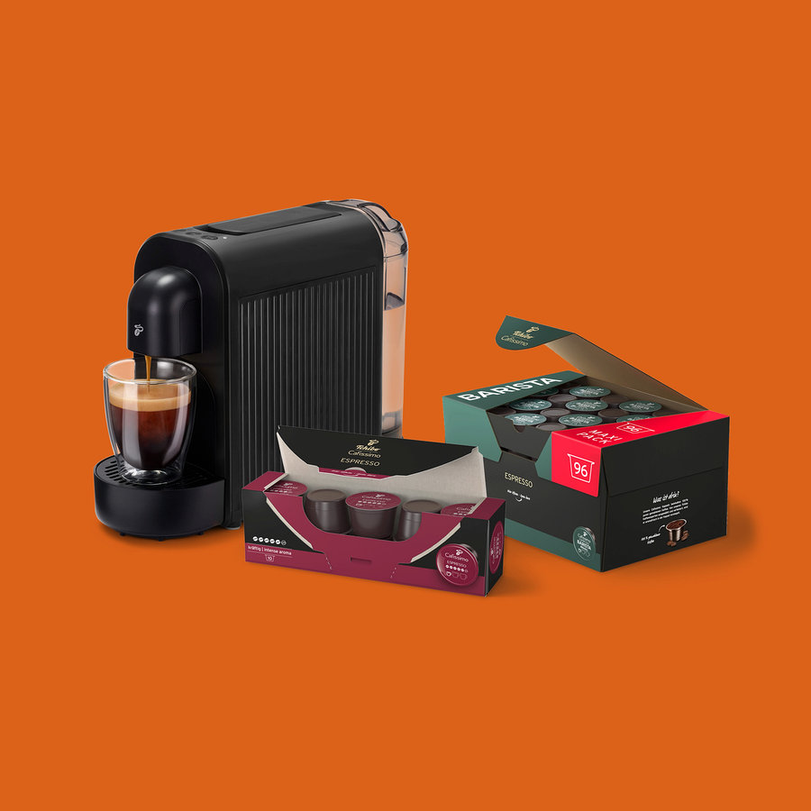 Fekete kávéfőző önt espressót egy üvegcsészébe, mellette egy nyitott doboz 96 Barista Espresso kapszulával és egy kisebb doboz Tchibo Cafissimo Espresso kapszulával.
