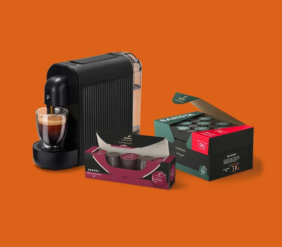 Fekete kávéfőző önt espressót egy üvegcsészébe, mellette egy nyitott doboz 96 Barista Espresso kapszulával és egy kisebb doboz Tchibo Cafissimo Espresso kapszulával.