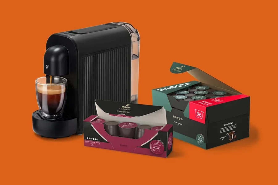 Fekete kávéfőző önt espressót egy üvegcsészébe, mellette egy nyitott doboz 96 Barista Espresso kapszulával és egy kisebb doboz Tchibo Cafissimo Espresso kapszulával.