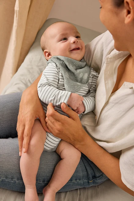 Anya tart egy mosolygó babát, aki állítható hosszúságú bodyt és muszlin kendőt visel.
