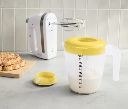 A pulton egy fehér mixer, egy sárga fedővel ellátott mérőpohár folyadékkal és egy halom gofri egy deszkán.