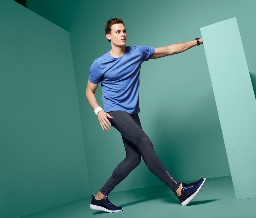 Férfi kék pólóban és sötétkék futónadrágban pózol egy zöld fal mellett.