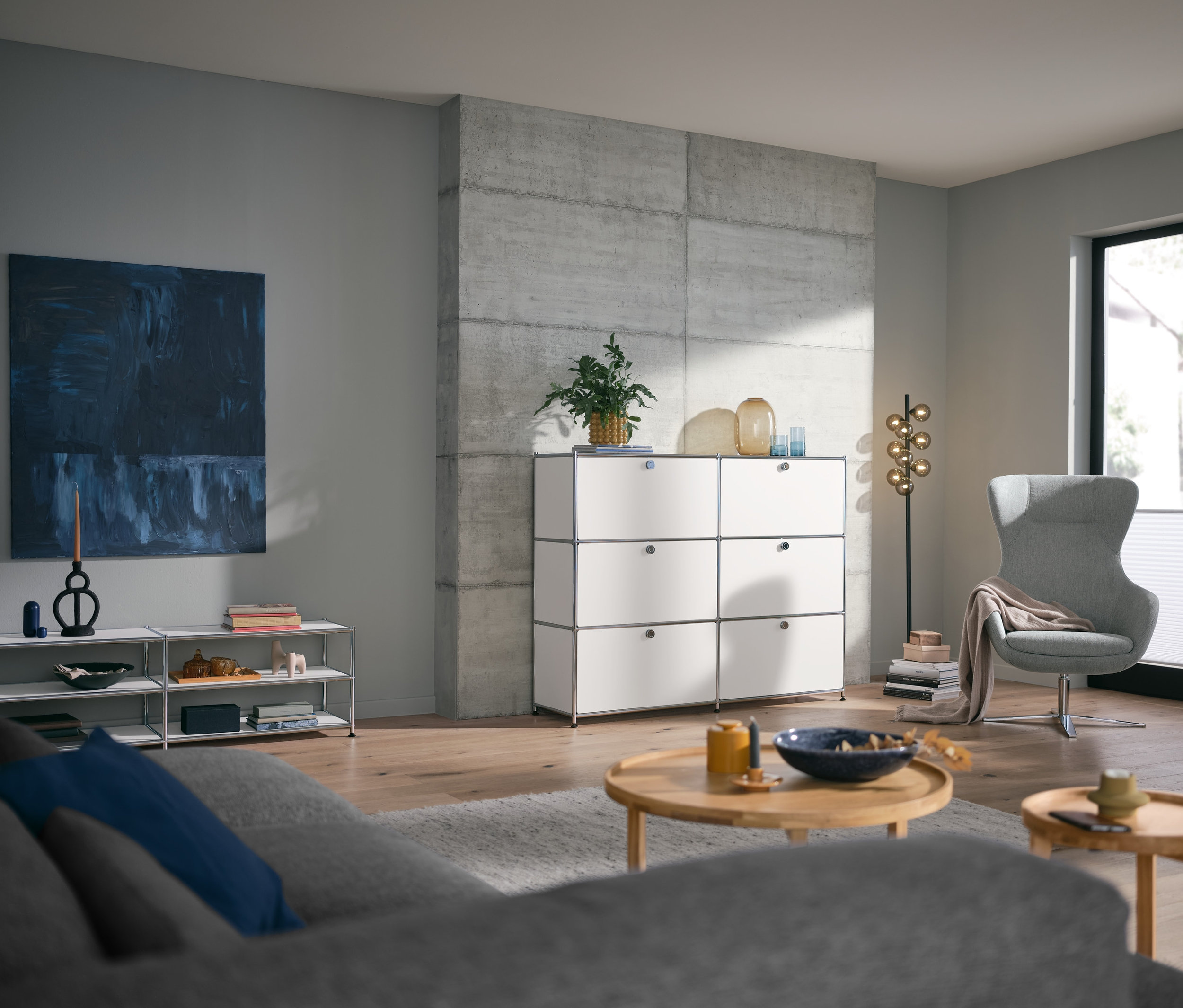 Nappali kerek dohányzóasztallal, fehér fém TV állvánnyal és 3x2 ajtós fém komóddal.