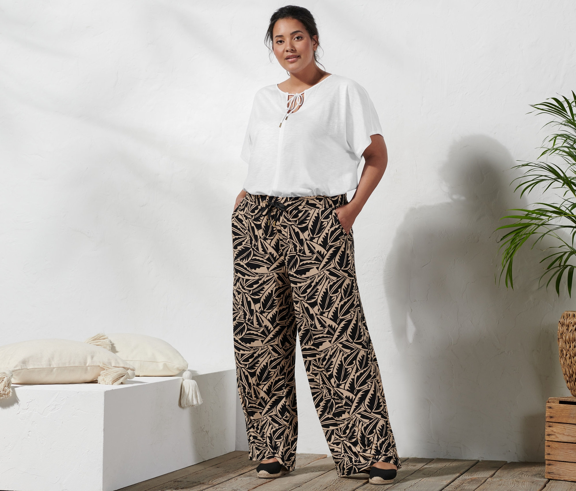 Egy nő áll, kezével a fekete-bézs mintás, női bőszárú nadrág zsebében, visel egy női denevérujjú pólóblúzt és egy fekete, magas sarkú szandált.