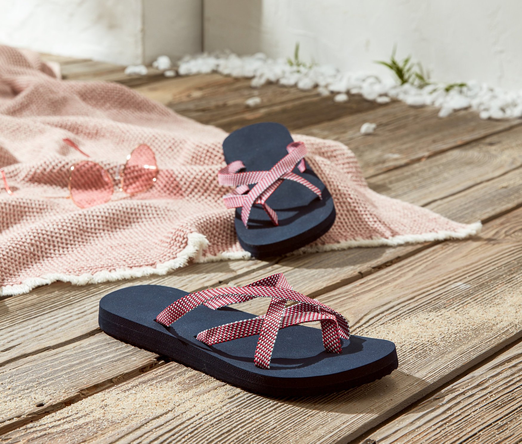 Két női lábujjas papucs egy fa padlón, rózsaszín napszemüveg és egy rózsaszín strandtakaró mellett.