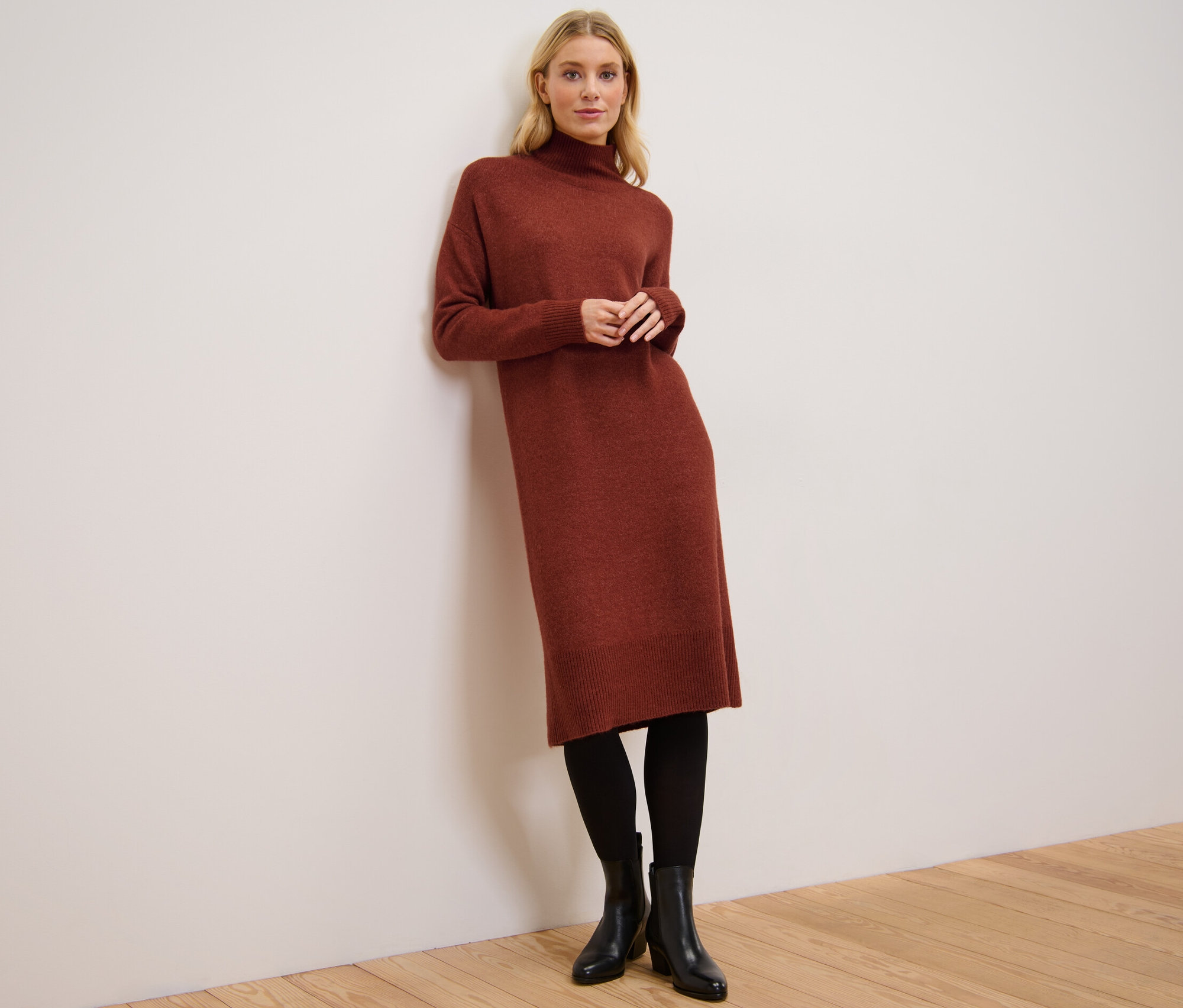 A falnak támaszkodó nő kötött ruhát visel gyapjúval, fekete harisnyát és bokacsizmát.