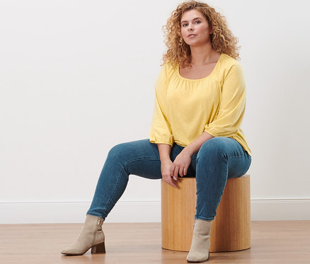 Egy nő ül egy fa zsámolyon, sárga, 3/4-es ujjú pólóblúzt és farmer hatású treggingst visel.
