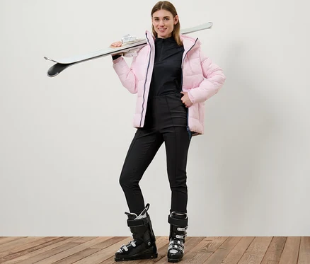 Nő pózol fekete női softshell sínadrágban és síbakancsban, a vállán sílécekkel.