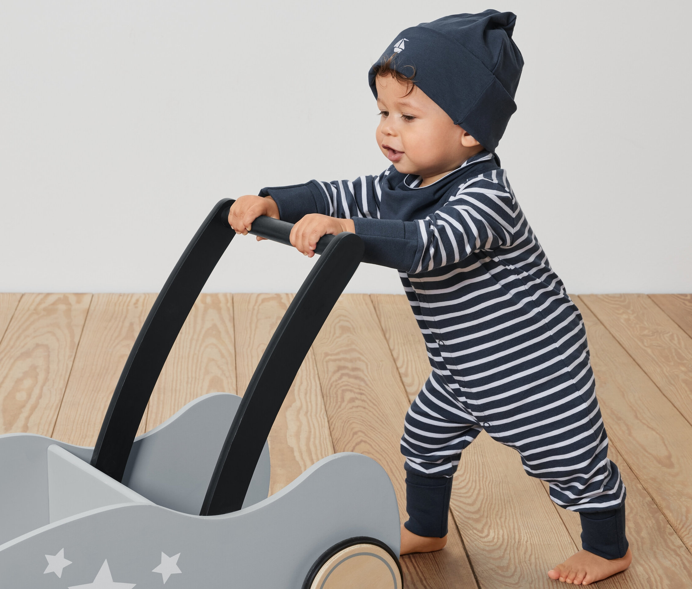 Kisgyermek tol egy szürke, csillagokkal díszített kocsit. Háromrészes együttes kisgyerekeknek van rajta: egy csíkos overall, egy sapka és egy előke.