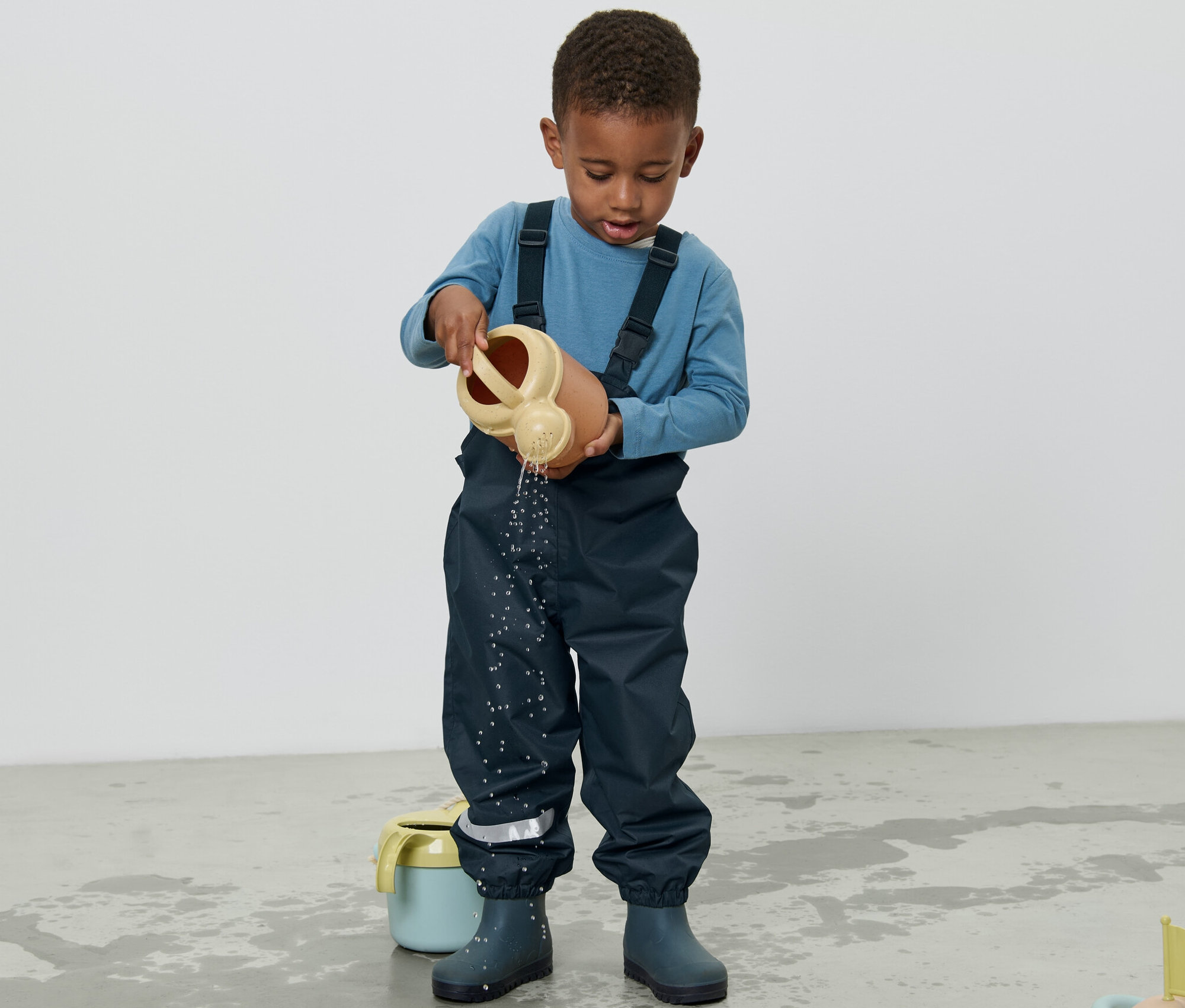 Egy kisfiú sötétkék kantáros esőnadrágban vizet önt a földre egy locsolókannából.