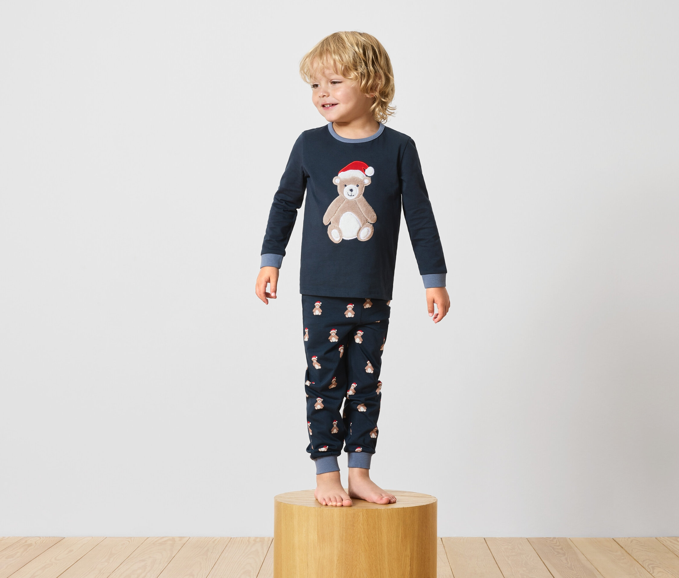 Egy fiú egy fa talapzaton áll, sötétkék, macimintás pizsamában.