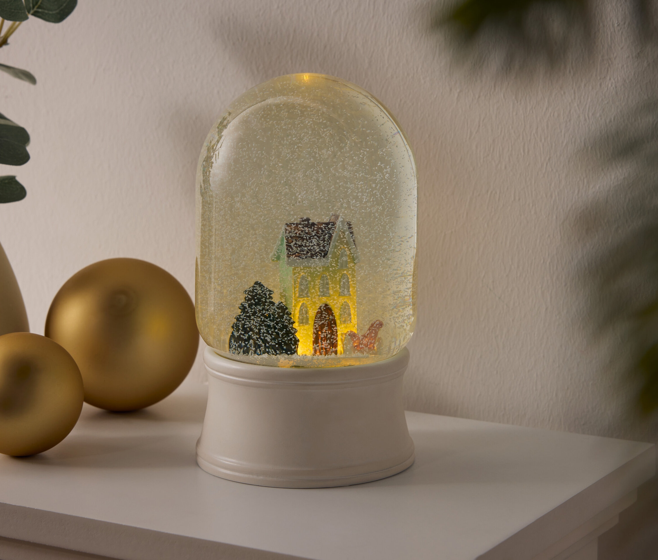 Hógömb házikóval és fákkal egy fehér talapzaton, egy polcon áll arany gömbök és egy növényekkel teli váza mellett.
