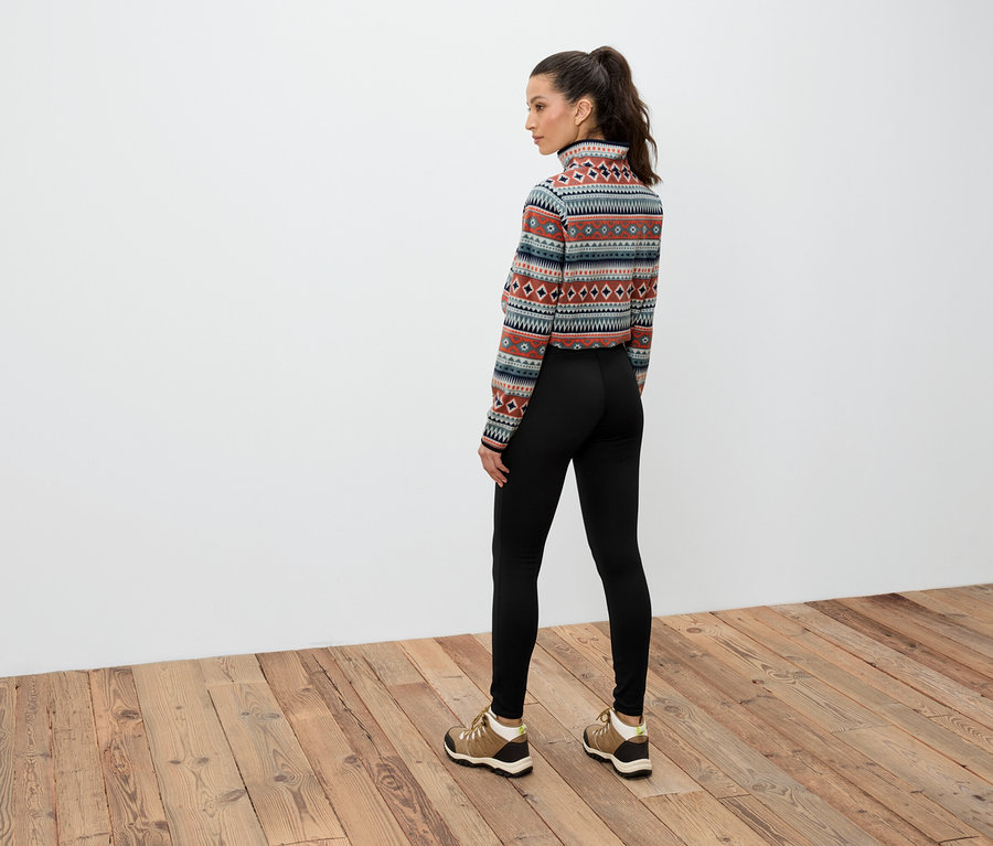 Egy barna hajú, lófarokba kötött nő áll háttal, mintás, hosszú ujjú felsőt, fekete leggingset és barna cipőt visel.