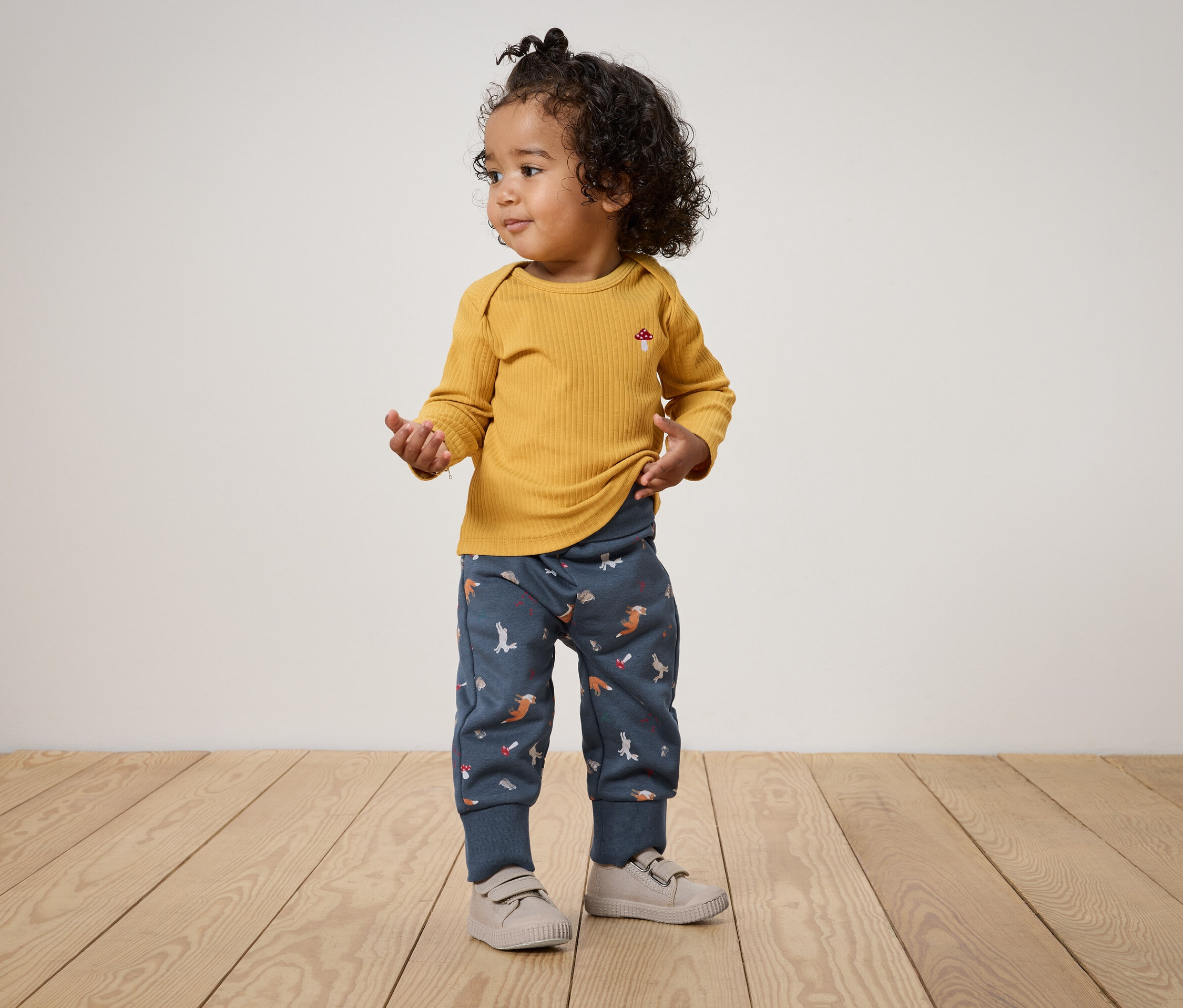 Egy göndör hajú gyermek áll egy fapadlón, mustársárga hosszú ujjú inget és kék, állatmintás nadrágot visel.