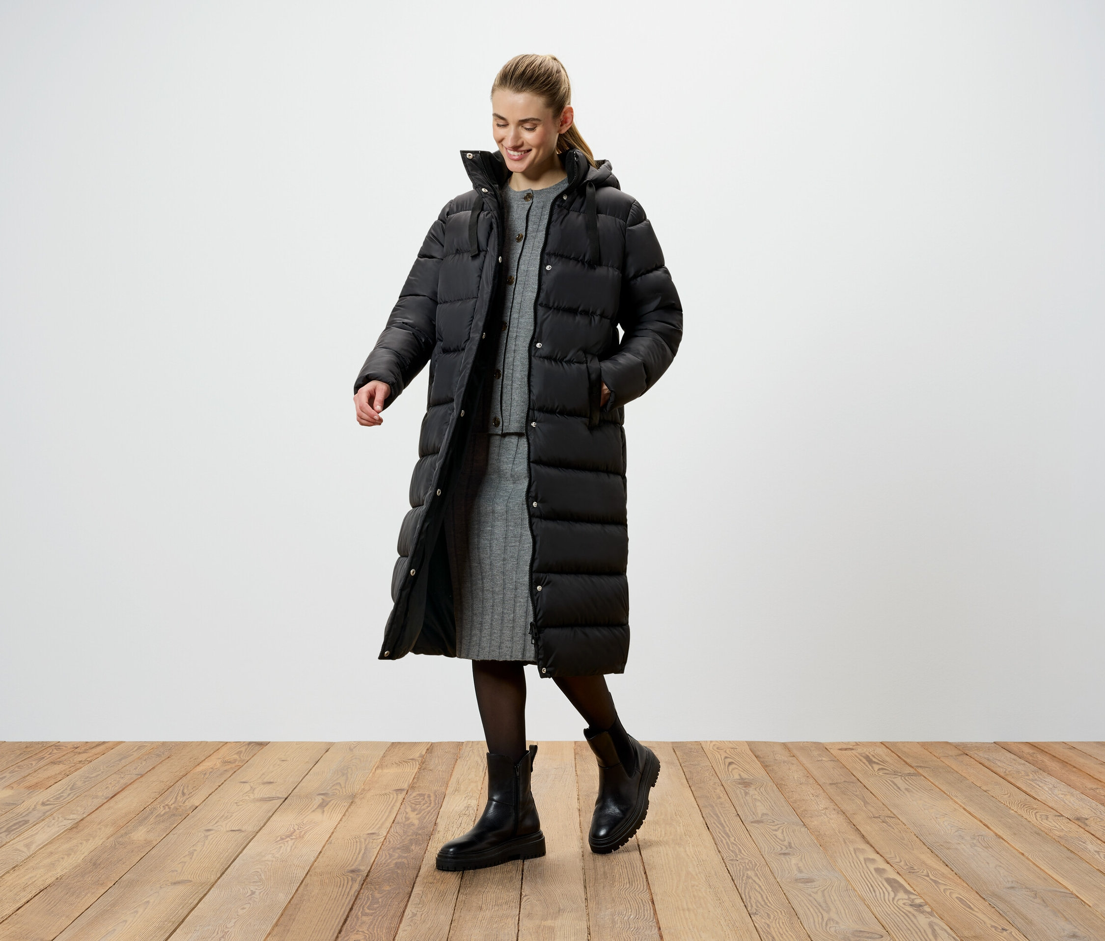 Egy nő hosszú, fekete, tollkabátban, szürke kötött ruhában és fekete csizmában áll egy fapadlón.