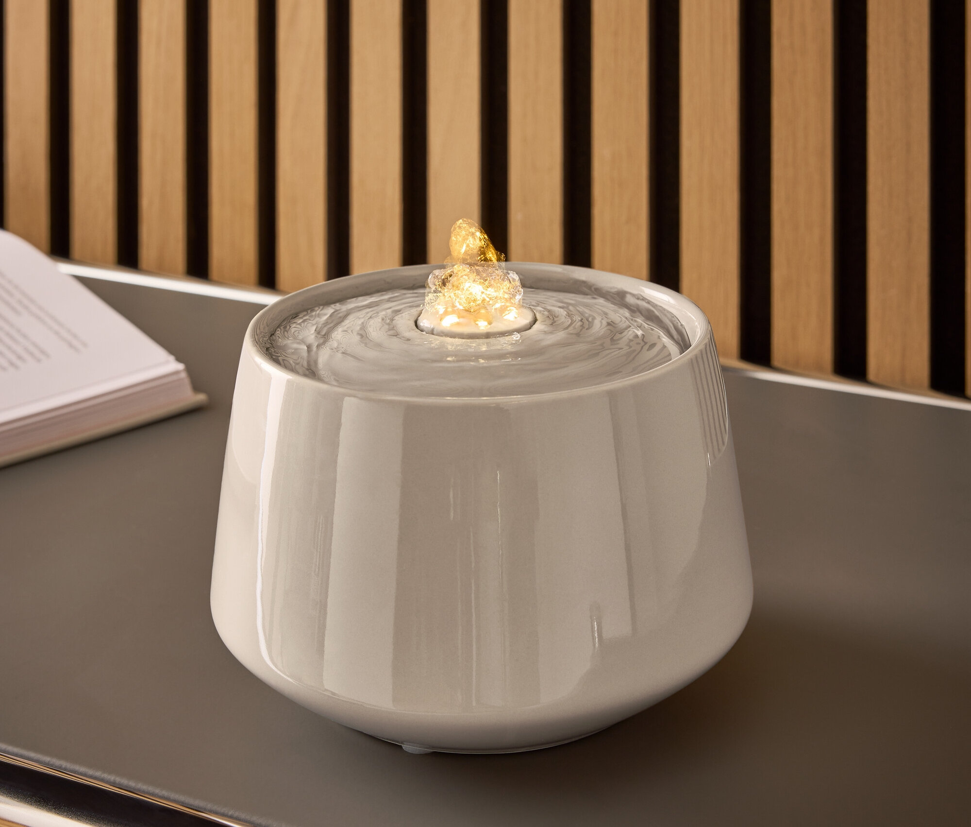 Szürke kerámia szökőkút vízzel és megvilágított kővel egy szürke asztalon.