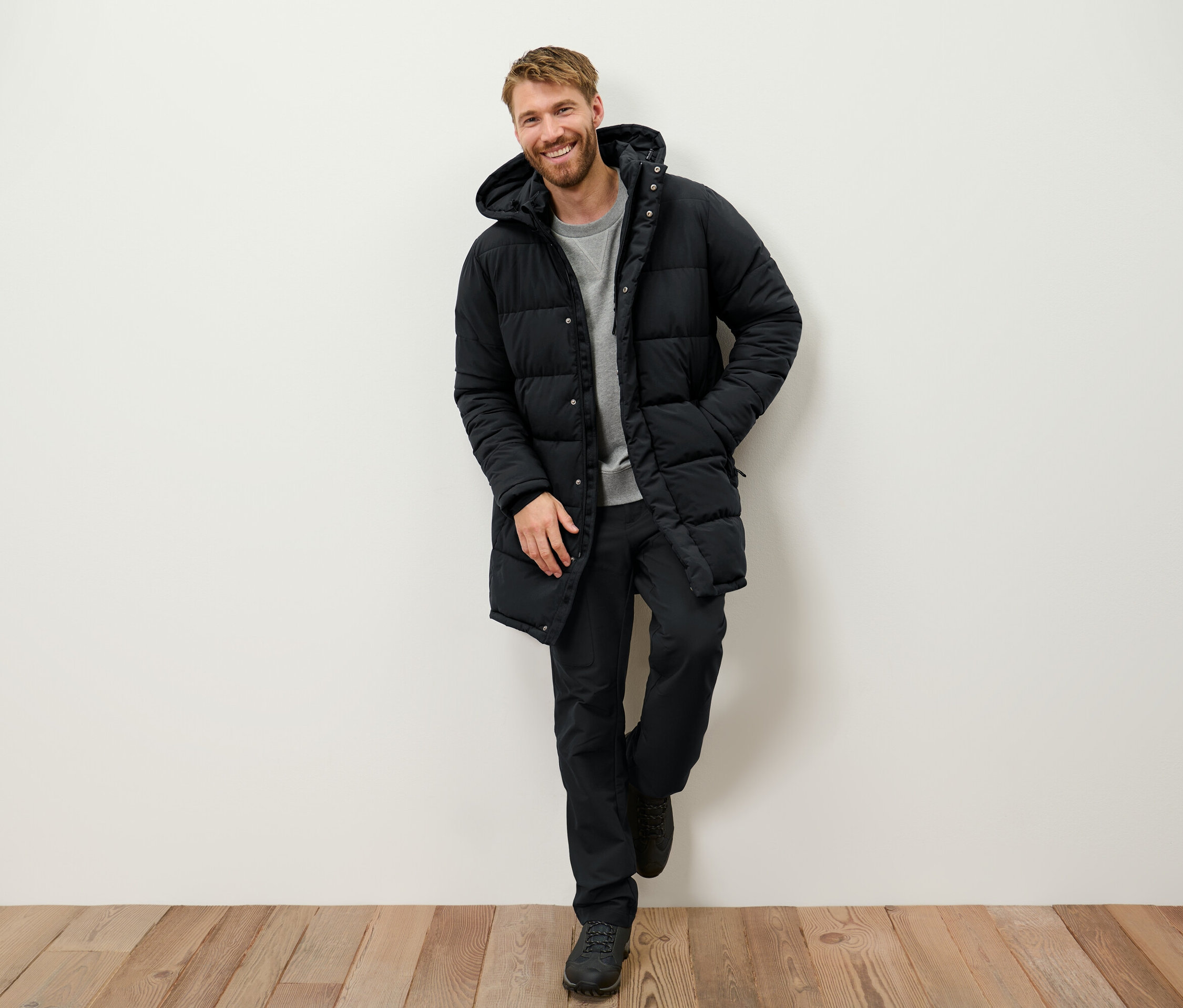 Egy mosolygó, szakállas férfi falnak támaszkodva fekete, kapucnis pehelykabátot, fekete nadrágot és csizmát visel.