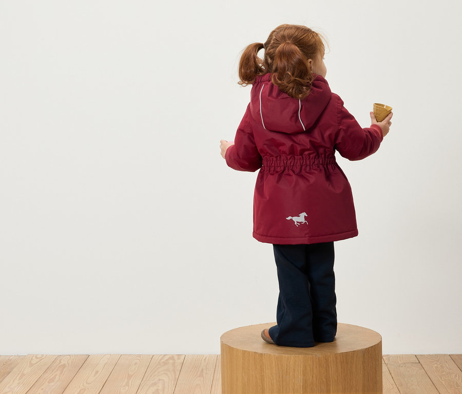 Egy vörös hajú, copfos kislány áll háttal a kamerának egy fa talapzaton, bordó színű, kapucnis kabátot visel, melyen egy ló képe van, és egy tölcsért tart a kezében.