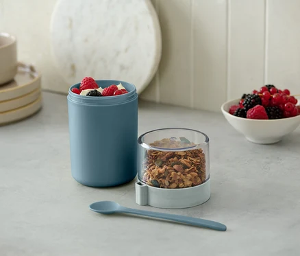 Egy zöld színű to-go joghurtos pohár müzlivel és bogyós gyümölcsökkel.