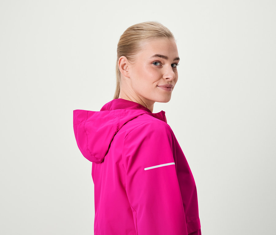 Szőke nő visszanéz a válla fölött, rózsaszín kapucnis kabátot visel.