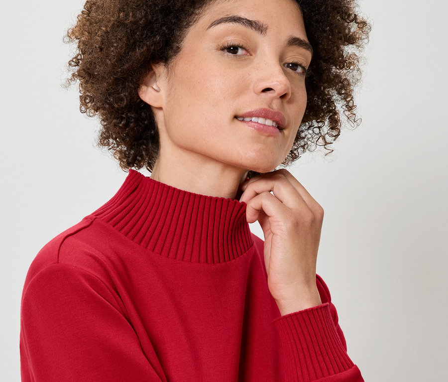 Egy göndör hajú nő portréja piros garbóban, kezét a nyakán tartja.