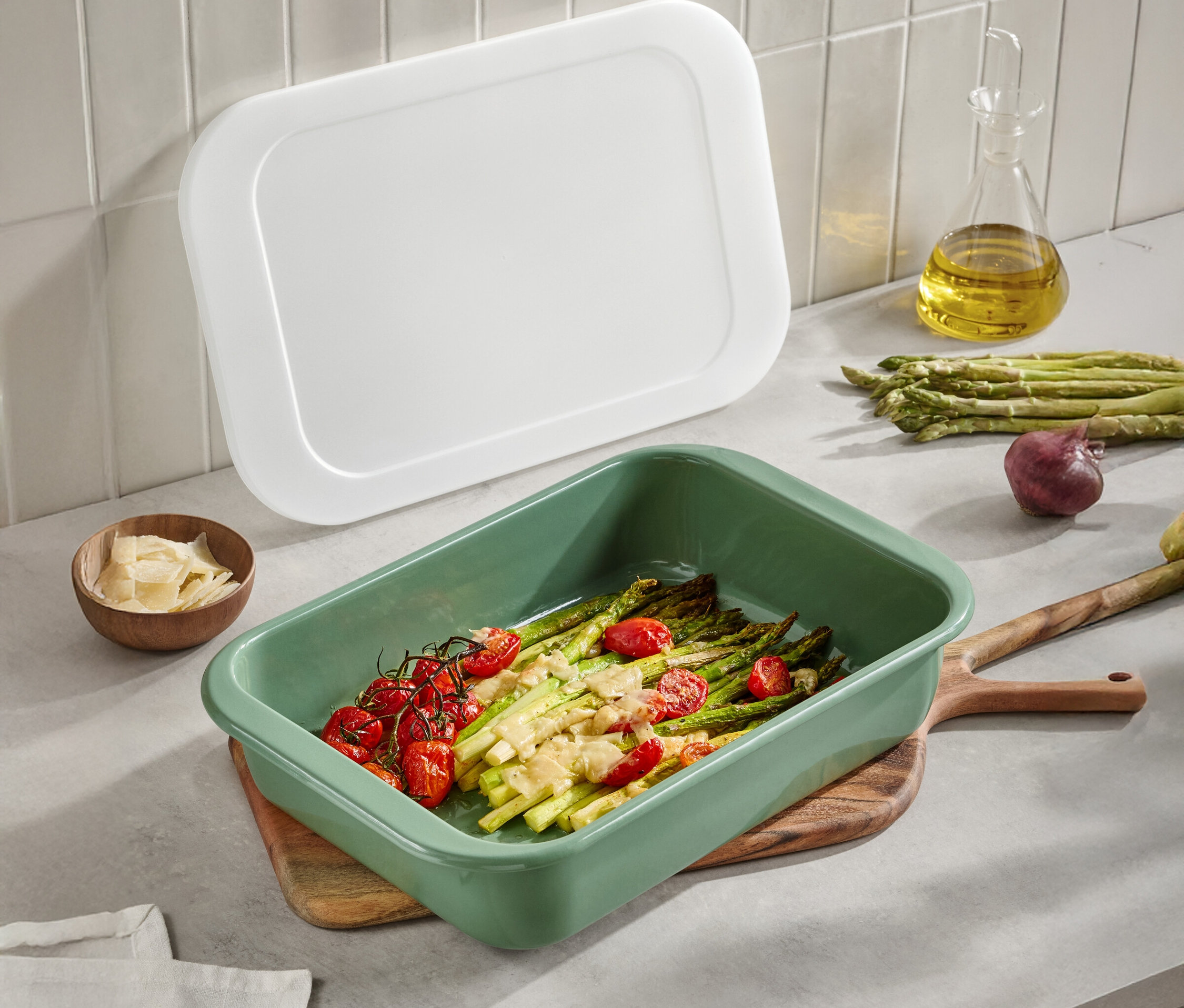 Zöld rakott edény spárgával és paradicsommal egy fa asztalon. Egy fehér fedő, olívaolaj, parmezán és hagyma is látható.