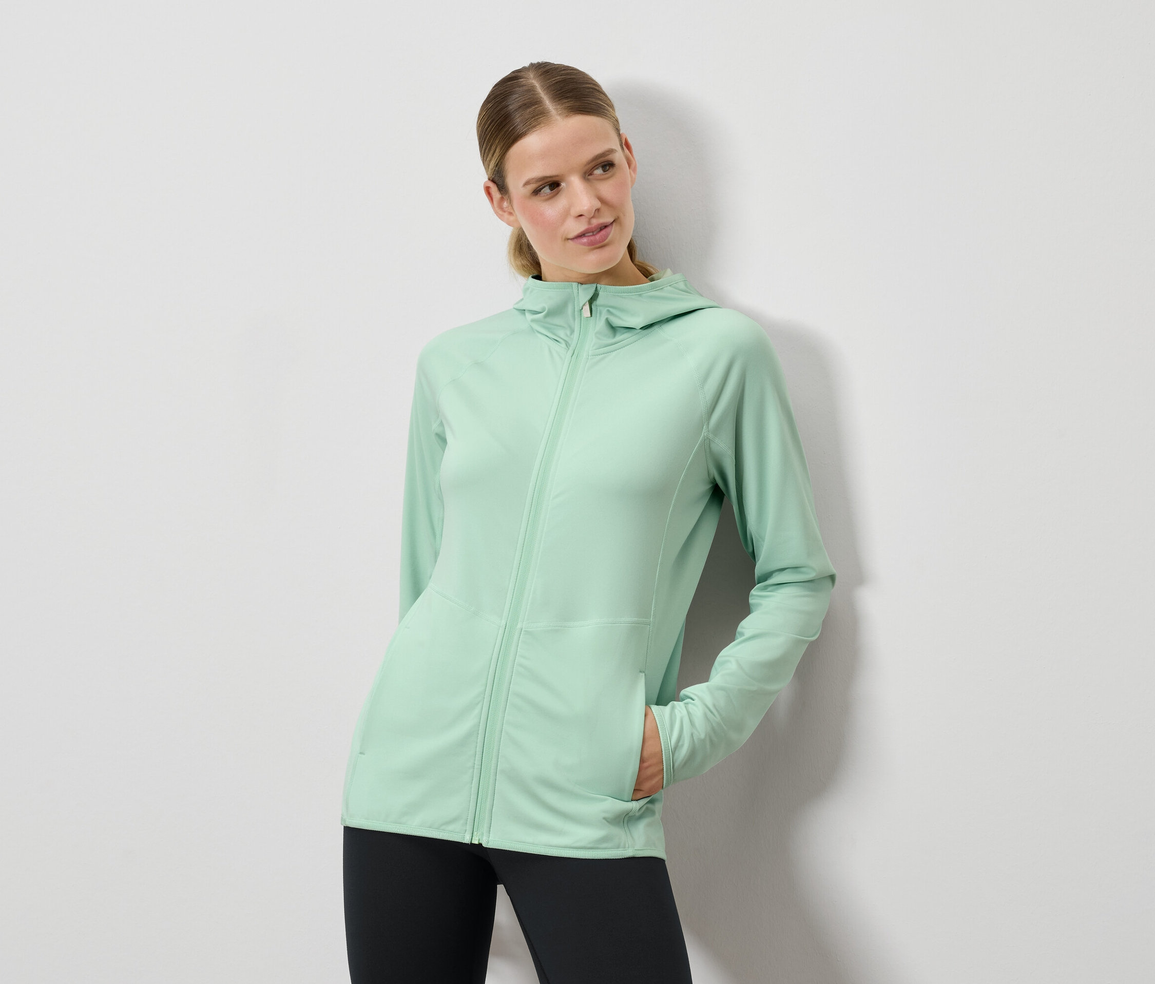 Nő pózol mentazöld kapucnis pulóverben és fekete leggingsben.