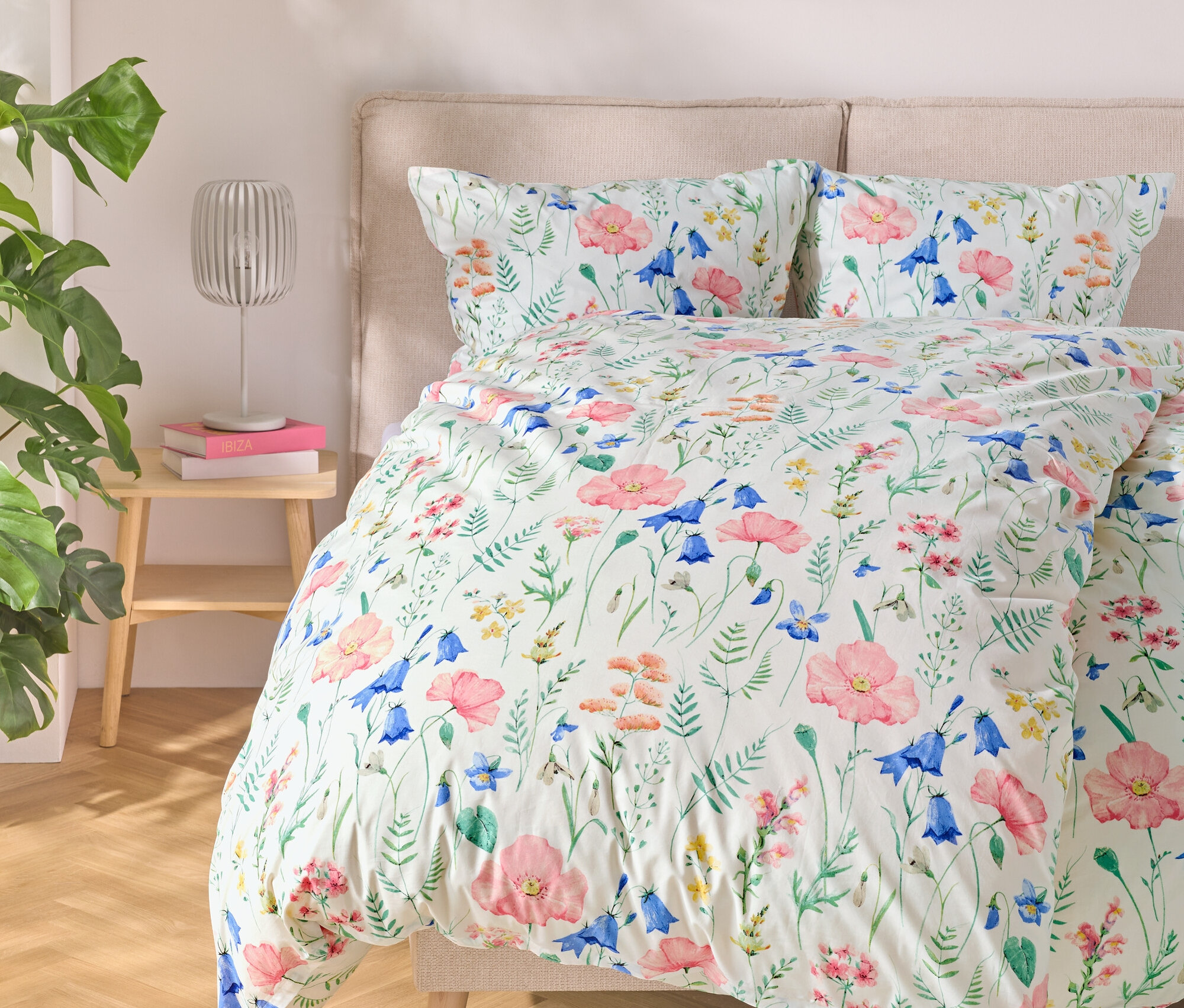 Virágmintás ágyneművel ellátott ágy, mellette asztal lámpával és könyvekkel, balra egy növény.