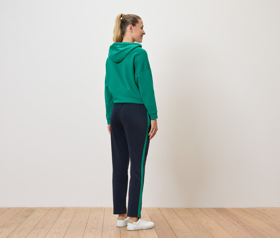 Egy nő háttal áll a kamerának, zöld kapucnis pulóvert és sötétkék, csíkos nadrágot visel.