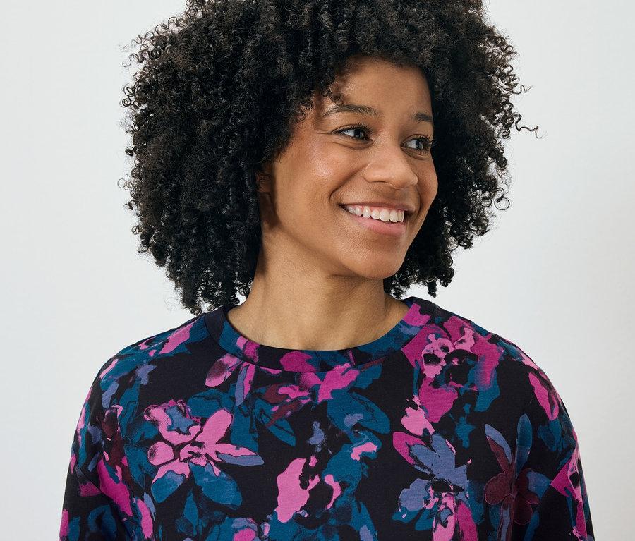 Portré egy mosolygó, afro frizurás nőről, aki rózsaszín és kék virágmintás fekete felsőt visel.