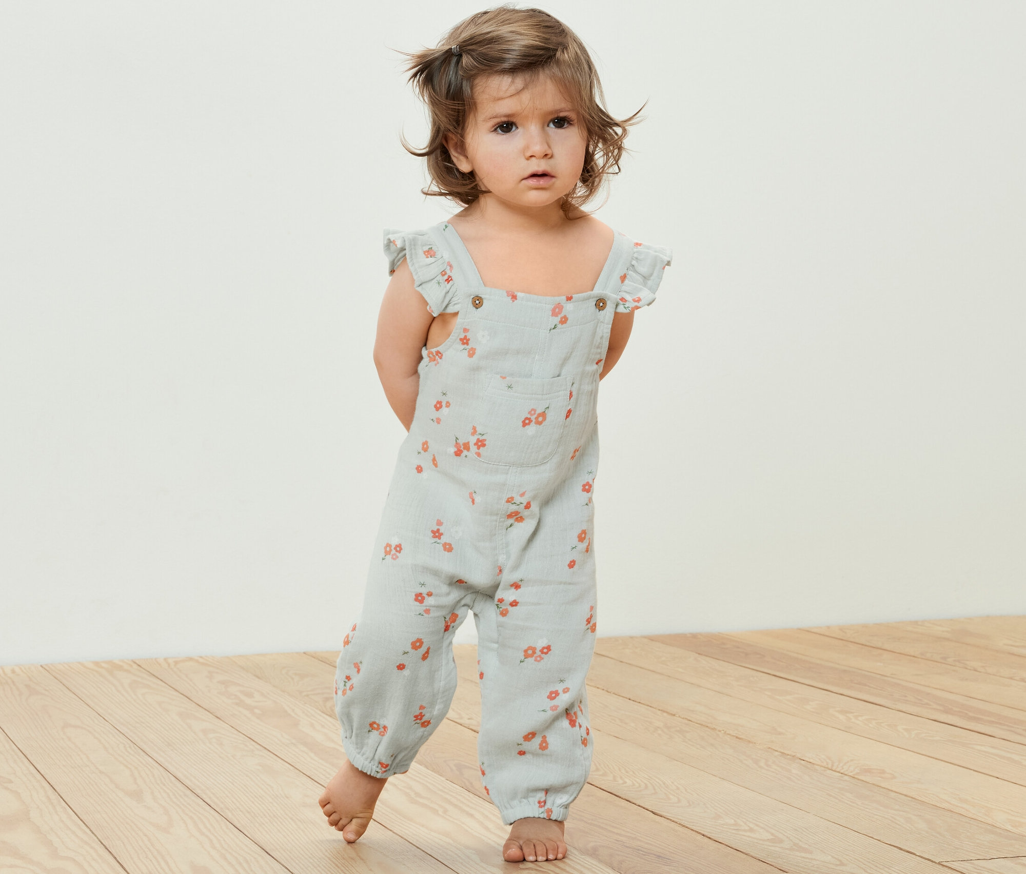 Egy kisgyermek áll egy fapadlón, virágmintás muszlin kezeslábast visel.