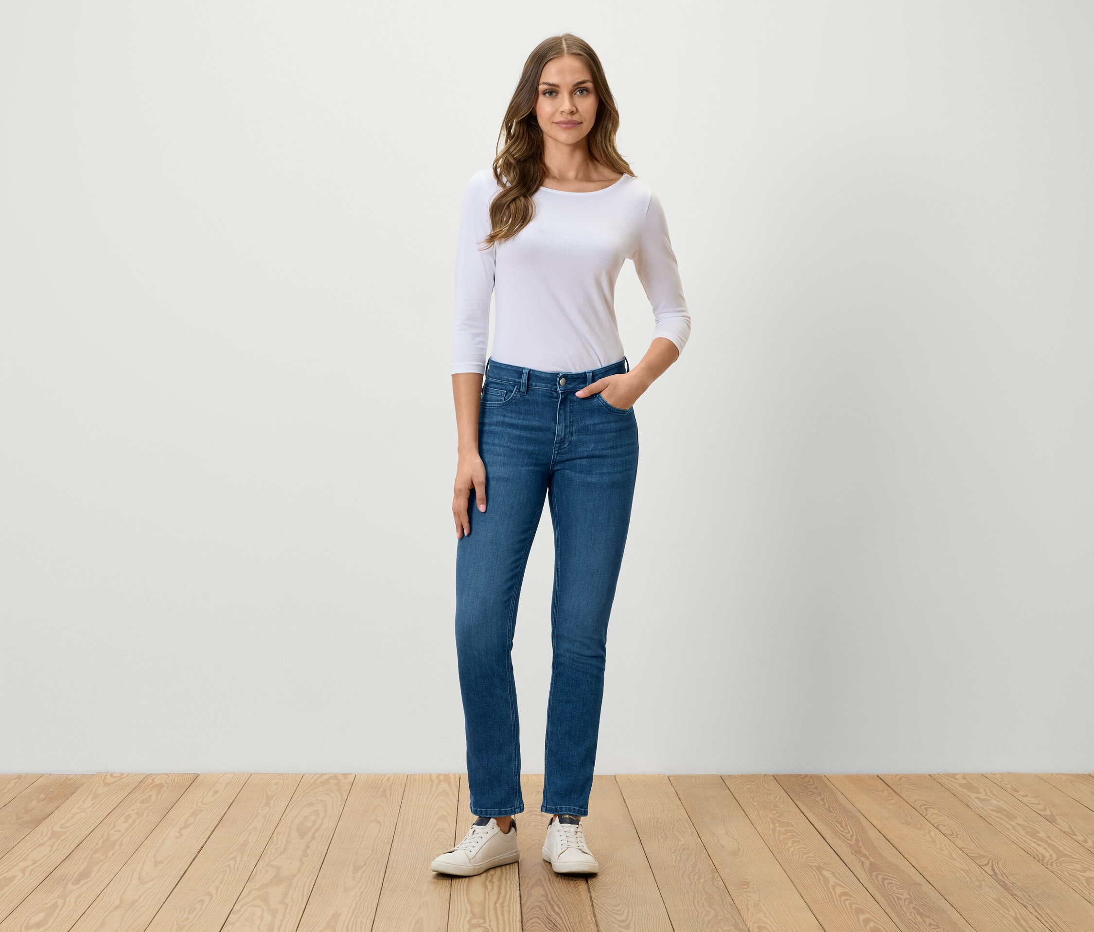 Egy nő áll és pózol kék farmerben, fehér pólóban és fehér tornacipőben.