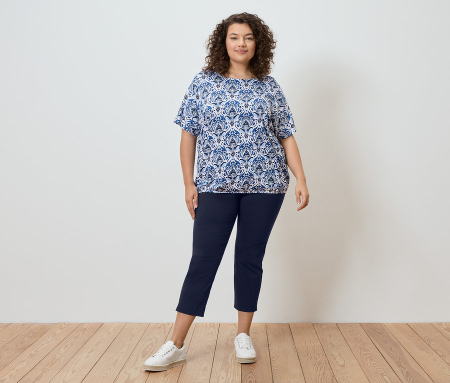 Egy göndör hajú nő áll egy fal előtt, kék mintás blúzt, sötétkék nadrágot és fehér tornacipőt visel.