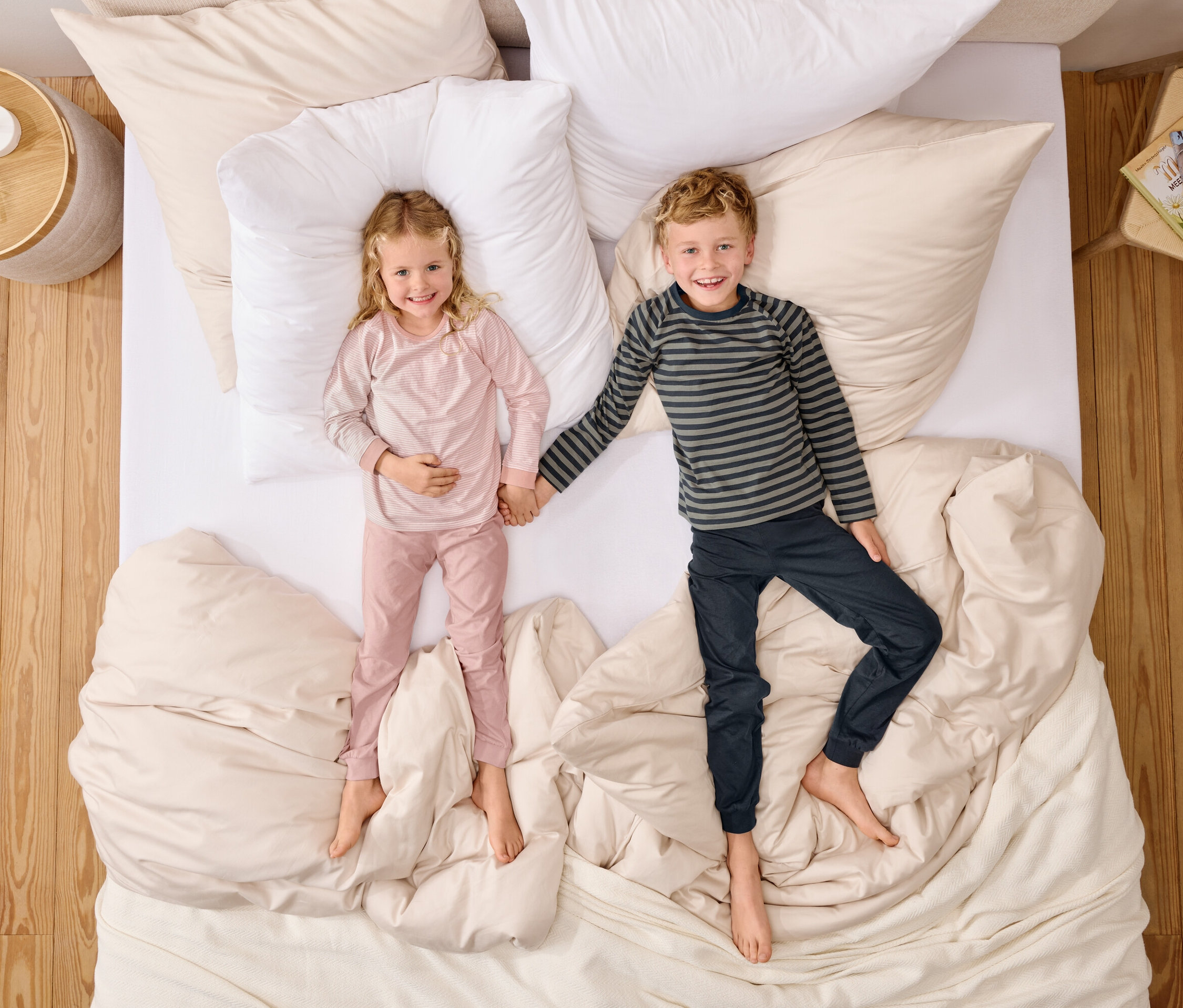 Felülnézet egy rózsaszín pizsamás lányról és egy csíkos felsős fiúról, akik kézen fogva fekszenek egy ágyon, fehér párnákkal.